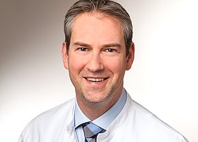 Dr. med. univ. Mario Reis Das Bild zeigt einen lächelnden Mann in einem weißen Arztkittel. Er hat kurzes, graues Haar und trägt ein hellblaues Hemd mit einer karierten Krawatte. Der Hintergrund ist hell und neutral.