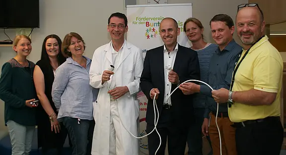 In einem Raum stehen mehrere Personen, darunter ein Arzt in weißem Kittel. Alle halten Kabel und Stecker in den Händen. Im Hintergrund ist ein Banner mit dem Schriftzug „Förderverein für den bunten Kreis“ zu sehen. Die Gruppe wirkt freundlich und engagiert.