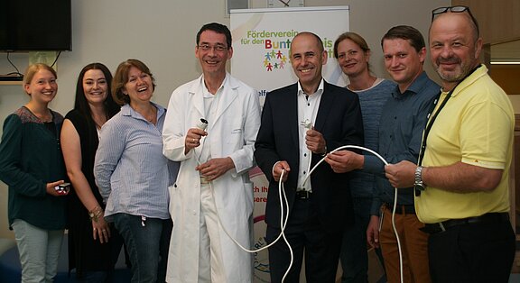 In einem Raum stehen mehrere Personen, darunter ein Arzt in weißem Kittel. Alle halten Kabel und Stecker in den Händen. Im Hintergrund ist ein Banner mit dem Schriftzug „Förderverein für den bunten Kreis“ zu sehen. Die Gruppe wirkt freundlich und engagiert.