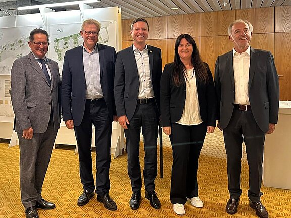 Fünf Erwachsene in Businesskleidung posieren lächelnd für ein Gruppenfoto. Sie stehen in einem Konferenzraum vor weißen Ausstellungstafeln mit Karten und Diagrammen, vermutlich zu einem Planungs- oder Architekturprojekt.