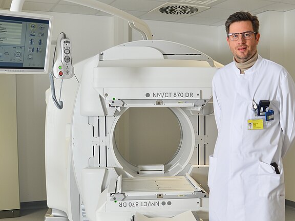 Ein Arzt in einem weißen Kittel steht neben einem modernen CT-Gerät. Das Gerät hat einen runden Öffnungsbereich und eine digitale Steuerung. Der Raum ist hell und klinisch. Der Arzt trägt eine Personalausweis-Karte und lächelt freundlich.
