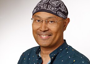 Ein Mann mit Brille und einem schwarzen Bandana lächelt in die Kamera. Er trägt ein dunkelblaues Hemd mit bunten Mustern. Der Hintergrund ist hell und einfarbig, was den Fokus auf sein Gesicht lenkt.