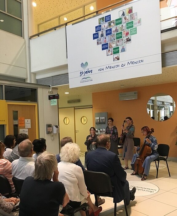 Kleines Streichensemble spielt vor sitzendem Publikum in einem Foyer; darüber ein Banner mit Herz aus Fotos und dem Slogan „30 Jahre – von Mensch zu Mensch“.
