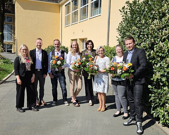 Eine Gruppe von sieben Personen steht im Freien vor einem gelben Gebäude. Sie halten bunte Blumensträuße in den Händen und lächeln. Die Szene vermittelt eine fröhliche und festliche Stimmung.