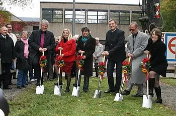 Eine Gruppe von fünf Personen steht mit Schaufeln in der Hand in einer Gartenlandschaft, bereit, einen Spatenstich zu vollziehen. Sie tragen festliche Kleidung und halten Blumensträuße. Im Hintergrund sind Zuschauer und ein Gebäude zu sehen, was auf eine öffentliche Veranstaltung hinweist.
