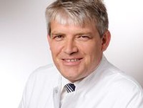 Dr. med Walter Aimer Ein lächelnder Arzt trägt einen weißen Kittel und eine Krawatte. Der Hintergrund ist hell und neutral. Die Person hat kurzes, graues Haar und blickt direkt in die Kamera.