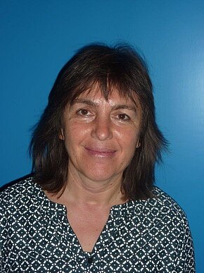 Porträtfoto einer Frau mit schulterlangem dunklen Haar. Sie lächelt und schaut direkt in die Kamera. Sie trägt ein schwarz-weiß gemustertes Oberteil; der Hintergrund ist einfarbig blau. Kopf- und Schulteranschnitt.