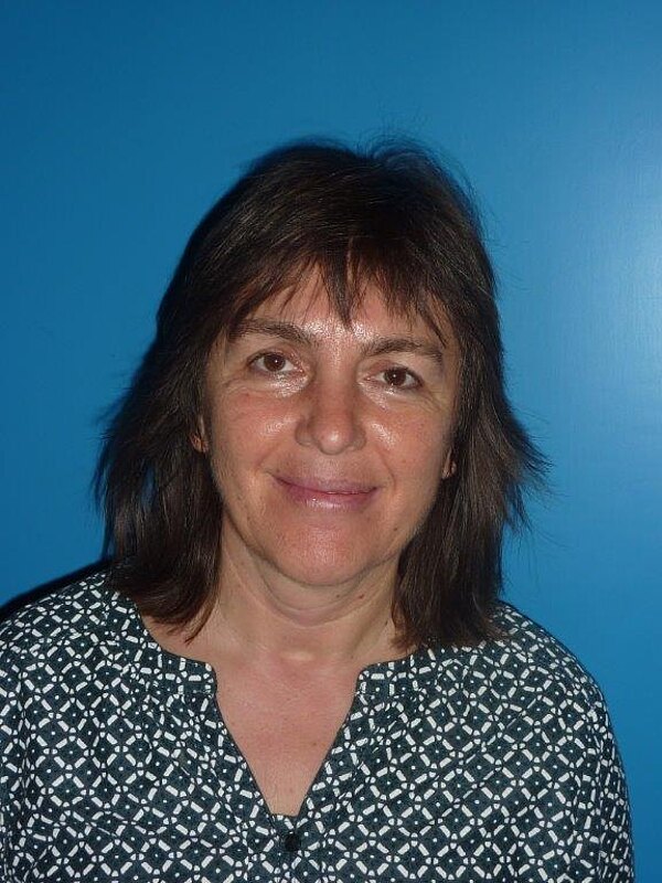 Porträtfoto einer Frau mit schulterlangem dunklen Haar. Sie lächelt und schaut direkt in die Kamera. Sie trägt ein schwarz-weiß gemustertes Oberteil; der Hintergrund ist einfarbig blau. Kopf- und Schulteranschnitt.