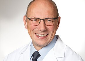 Porträt eines lächelnden, glatzköpfigen Mannes mit Brille in weißem Arztkittel, hellblauem Hemd und dunkler Krawatte vor neutralem Hintergrund.