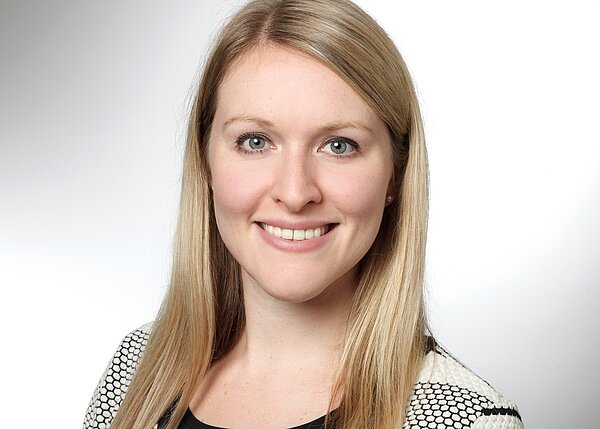 Porträt einer lächelnden Frau mit langen, blonden Haaren. Sie trägt ein schwarzes Oberteil und eine jackenähnliche Bluse mit einem grafischen Muster. Der Hintergrund ist hell und neutral, was den Fokus auf ihr Gesicht lenkt.