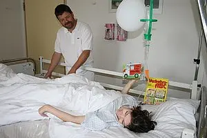Ein Pfleger steht an einem Krankenhausbett, in dem ein Kind mit einem Verband schläft. Das Kind hält ein Spielzeug-Lastwagen und ein Buch in der Hand. Im Hintergrund ist ein Tropf zu sehen, der über dem Bett hängt.