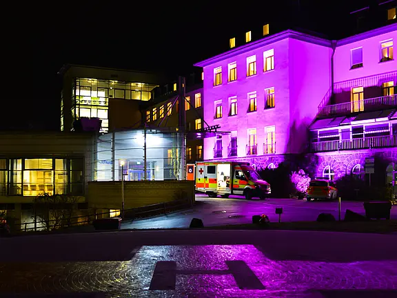 Nachtszene eines Krankenhauses: Ein mehrstöckiges Gebäude ist in violettem Licht beleuchtet, Fenster erstrahlen warm. Vor dem Eingang steht ein Rettungswagen. Im Vordergrund erkennbar ein beleuchteter Hubschrauberlandeplatz.