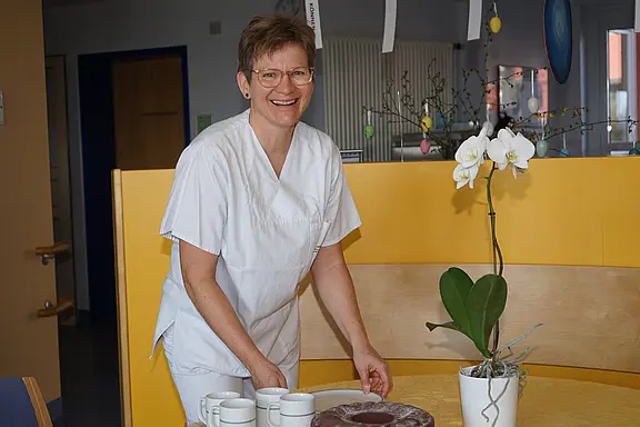 Eine lächelnde Pflegekraft in weißer Arbeitskleidung steht an einem Tisch und serviert Tassen. Neben ihr steht eine blühende Orchidee in einem weißen Topf. Die Umgebung ist hell und freundlich gestaltet.