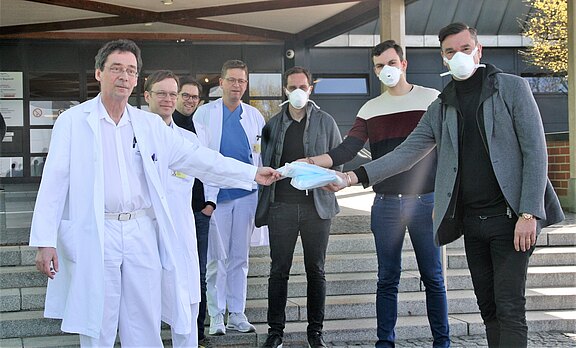 Vor einem Klinikgebäude stehen mehrere Personen, einige in weißer Arztkleidung. Zwei Männer mit Atemschutz reichen dem Arzt ein Paket OP-Masken, als symbolische Übergabe. Der Fokus liegt auf Schutzmaßnahmen und Zusammenarbeit im Gesundheitswesen.