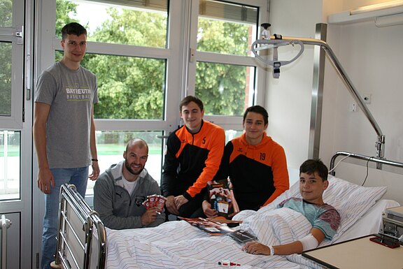 Im Krankenhauszimmer liegt ein Junge im Bett, lächelnd mit verbundenem Arm. Vier junge Männer, zwei in orange-schwarzen Trainingsjacken, besuchen ihn und überreichen Autogrammkarten. Die Gruppe lacht freundlich; es ist ein aufmunternder Krankenbesuch.