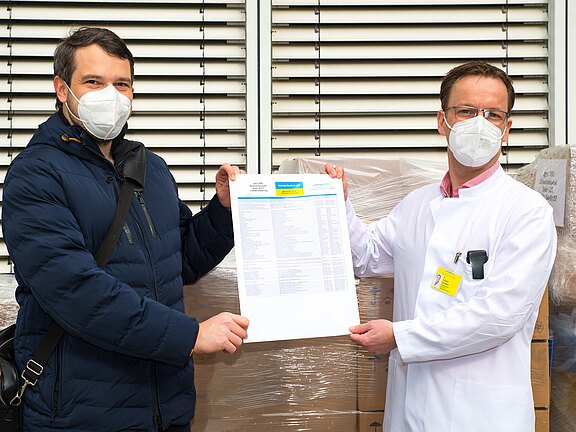 Zwei Personen mit Schutzmasken halten gemeinsam ein Dokument in die Kamera; eine trägt einen weißen Arztkittel. Im Hintergrund stehen verpackte Kartons. Die Szene wirkt wie eine Übergabe oder Bestätigung von Lieferware.