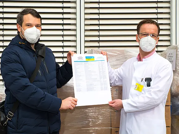 Zwei Personen mit Schutzmasken halten gemeinsam ein Dokument in die Kamera; eine trägt einen weißen Arztkittel. Im Hintergrund stehen verpackte Kartons. Die Szene wirkt wie eine Übergabe oder Bestätigung von Lieferware.