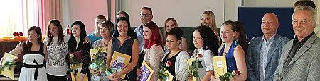 Eine Gruppe von Absolventen steht auf einer Bühne und hält ihre Zertifikate in den Händen. Einige von ihnen tragen Blumen. Es gibt auch einige Personen im Hintergrund, die die Absolventen unterstützen. Die Stimmung wirkt feierlich und erfreulich.