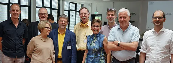 Gruppe von neun Erwachsenen steht in einem hellen Konferenzraum, lächelt in die Kamera. Einige tragen Namensschilder; Kleidung ist business-casual. Stimmung freundlich, wirkt wie ein Workshop oder Netzwerktreffen.