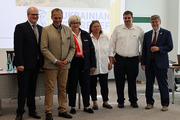 Sechs Erwachsene stehen nebeneinander in einem Konferenzraum. Zwei tragen Anzüge, vier Uniformen einer Hilfsorganisation. Sie blicken zur Kamera, neutral bis freundlich. Im Hintergrund eine Präsentationsfolie mit der Aufschrift „Ukrainian Relief“. Der Rahmen wirkt wie eine offizielle Veranstaltung oder Pressefoto.