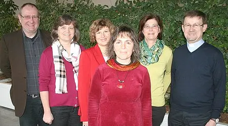 Gruppenfoto: Sechs Erwachsene stehen nebeneinander und lächeln in die Kamera, vor einer grünen Pflanzenwand. Freundliche, informelle Stimmung.