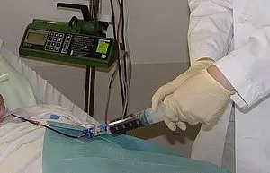 Eine medizinische Fachkraft in einem weißen Kittel und Handschuhen verabreicht mit einer Spritze eine Behandlung an einem Patienten. Der Patient liegt auf einer Liege, umgeben von medizinischen Geräten.