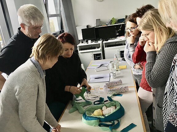 Mehrere Erwachsene stehen in einem Schulungsraum um einen Tisch. Eine Frau demonstriert an einer Babypuppe mit Tüchern das richtige Wickeln. Alle schauen konzentriert zu; Notizbücher liegen bereit.