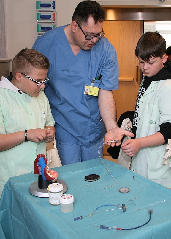 Ein Arzt in blauer Arbeitskleidung erklärt zwei Kindern, die in Ärzte-ähnlichen Kitteln gekleidet sind, medizinische Geräte und Modelle. Auf einem Tisch liegen verschiedene Utensilien und ein anatomisches Modell eines Herzens. Alle drei Personen zeigen Interesse und sind in das Geschehen konzentriert.