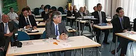 Eine Gruppe von Männern in Anzügen sitzt an Tischen in einem Konferenzraum. Sie hören aufmerksam zu, während eine Präsentation stattfindet. Im Hintergrund sind einige weitere Personen und ein Projektor sichtbar. Das Ambiente ist professionell und formell.