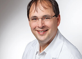 Dr. med. Klaus Lang-Schwarz Porträtfoto: Ein lächelnder Mann mit Brille trägt einen weißen Kittel und blickt in die Kamera vor neutralem Hintergrund.
