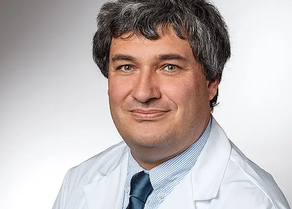 Porträt einer erwachsenen Person in weißem Kittel und dunkler Krawatte, leicht lächelnd, vor neutralem Hintergrund. Fokus auf Gesicht und professionelle Kleidung, ähnlich einem offiziellen Mitarbeiterfoto.