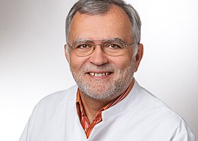 Prof. Dr. med. Robert Offner Porträt eines älteren Mannes mit grauen Haaren, Brille und kurzem Bart. Er lächelt freundlich und trägt einen weißen Kittel über einem orangegestreiften Hemd vor neutralem Hintergrund.
