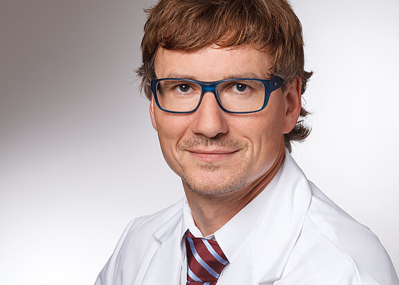 Porträt eines Mannes mit braunen Haaren und blauer Brille, der freundlich in die Kamera blickt. Er trägt einen weißen Kittel und eine rot-weiß gestreifte Krawatte vor neutralem Hintergrund.