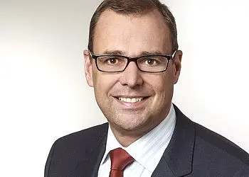 Porträt eines lächelnden Mannes mit Brille, im dunklen Anzug und gestreiftem Hemd mit roter Krawatte, vor hellem, neutralem Hintergrund.