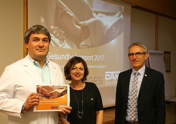 Drei Personen stehen vor einer Leinwand mit der Folie „Gesundheitsreport 2017“. Links hält eine Person im weißen Kittel das gedruckte Heft in die Kamera. Neben ihr stehen zwei weitere Personen in Businesskleidung. Die Szene wirkt wie eine Pressevorstellung oder Präsentation des Reports.