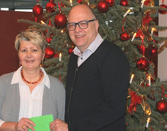 Zwei Personen stehen lächelnd vor einem Weihnachtsbaum, der mit roten Kugeln und goldenen Schleifen geschmückt ist. Eine Person hält einen grünen Umschlag in der Hand, während die andere Person daneben steht. Die Szene vermittelt eine festliche Stimmung.