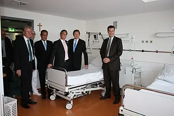Sechs Männer stehen in einem Krankenhauszimmer mit zwei leeren Betten. Sie tragen formelle Kleidung und lächeln. Im Hintergrund sind medizinische Geräte und helle Wände zu sehen, was eine moderne Krankenhausumgebung vermittelt.