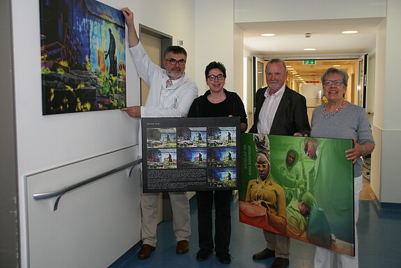 Vier Personen stehen in einem Flur und halten große Bilder sowie eine Fototafel. Einer der Männer hängt ein Bild an die Wand. Die Szene zeigt eine künstlerische Ausstellung oder Präsentation. Das Licht ist hell und der Raum wirkt freundlich.