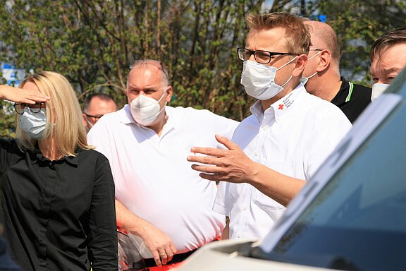 Eine Gruppe von Menschen diskutiert im Freien, alle tragen Masken. Im Vordergrund steht ein Mann in einem weißen Hemd mit dem Logo des Deutschen Roten Kreuzes und erklärt etwas, während eine Frau ihm aufmerksam zuhört und sich die Augen mit der Hand schützt.