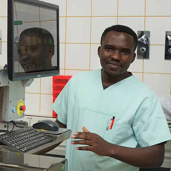 Medizinischer Mitarbeiter in mintgrüner OP-Kleidung steht an einem Computerarbeitsplatz im Krankenhaus und bedient Tastatur und Maus; sterile Kacheln im Hintergrund deuten auf Klinik- oder Laborumgebung hin.