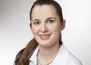 Angelika Pastor Ein Porträt einer lächelnden Frau mit langen, braunen Haaren, die in einem weißen Hemd und einer Perlenkette gekleidet ist. Sie blickt direkt in die Kamera, mit freundlichen, blauen Augen vor einem hellen Hintergrund.