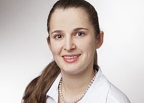 Angelika Pastor Ein Porträt einer lächelnden Frau mit langen, braunen Haaren, die in einem weißen Hemd und einer Perlenkette gekleidet ist. Sie blickt direkt in die Kamera, mit freundlichen, blauen Augen vor einem hellen Hintergrund.