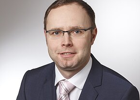 Thomas Haas Porträtfoto: Ein Mann mit kurzer Frisur und Brille trägt dunklen Anzug, weißes Hemd und hell gestreifte Krawatte. Er blickt in die Kamera und lächelt leicht vor neutralem Hintergrund.