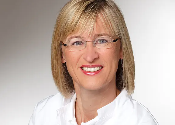 Porträtfoto: Lächelnde erwachsene Person mit schulterlangem blondem Haar und Brille, blickt direkt in die Kamera. Trägt ein weißes Oberteil vor neutralem, hellgrauem Hintergrund.