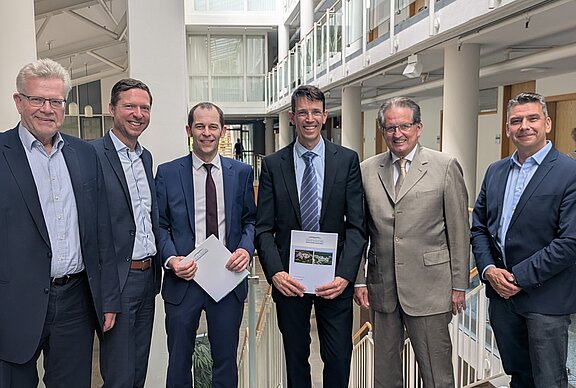 Sechs Personen stehen in einem modernen Bürogebäude, lächeln und halten Dokumente in den Händen. Sie sind formell gekleidet und strahlen eine positive Atmosphäre aus. Im Hintergrund sind helle, offene Räumlichkeiten mit Glaswänden und Treppen zu sehen.