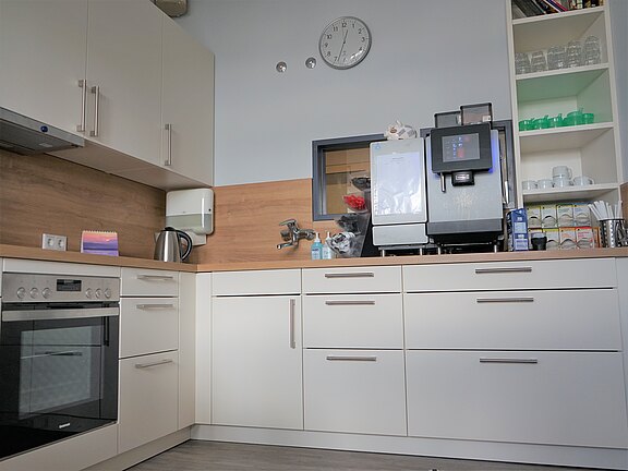 Moderne Teeküche mit weißen Schränken und Holzarbeitsplatte: links Einbauherd, mittig Spüle, rechts große Kaffeemaschine. Daneben offene Regale mit Gläsern und Tassen. Ordentlich und hell, wirkt wie eine Büroküche.