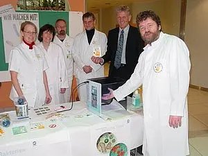 Sechs Personen in weißen Kitteln stehen um einen Tisch mit Informationsmaterialien. Einer der Männer zeigt auf einen Bildschirm. Im Hintergrund ist ein Plakat mit der Aufschrift „WIR MACHEN MIT!“ zu sehen. Die Personen wirken engagiert und freundlich.