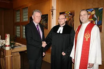 Drei Männer stehen in einem Raum mit religiösem Hintergrund. Einer trägt einen schwarzen Talar und begrüßt einen anderen im Anzug mit einem Handschlag. Der dritte Mann ist in liturgischer Kleidung gekleidet. Die Atmosphäre wirkt feierlich und freundlich.
