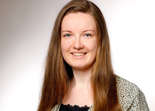 Porträt einer lächelnden Frau mit langen braunen Haaren, direkter Blick in die Kamera, heller neutraler Hintergrund; professionelle Profilaufnahme.