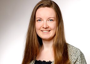 Porträt einer lächelnden Frau mit langen braunen Haaren, direkter Blick in die Kamera, heller neutraler Hintergrund; professionelle Profilaufnahme.